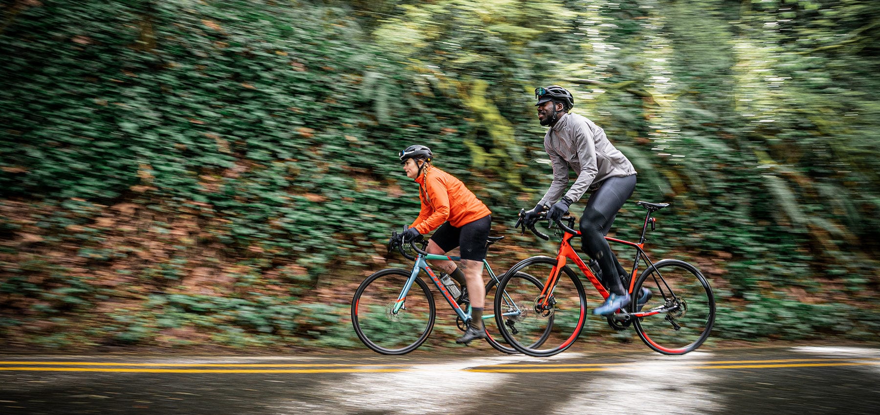 A man and woman riding side by side on  road bikes in the rain, wearing the Cloudburst waterproof jacket