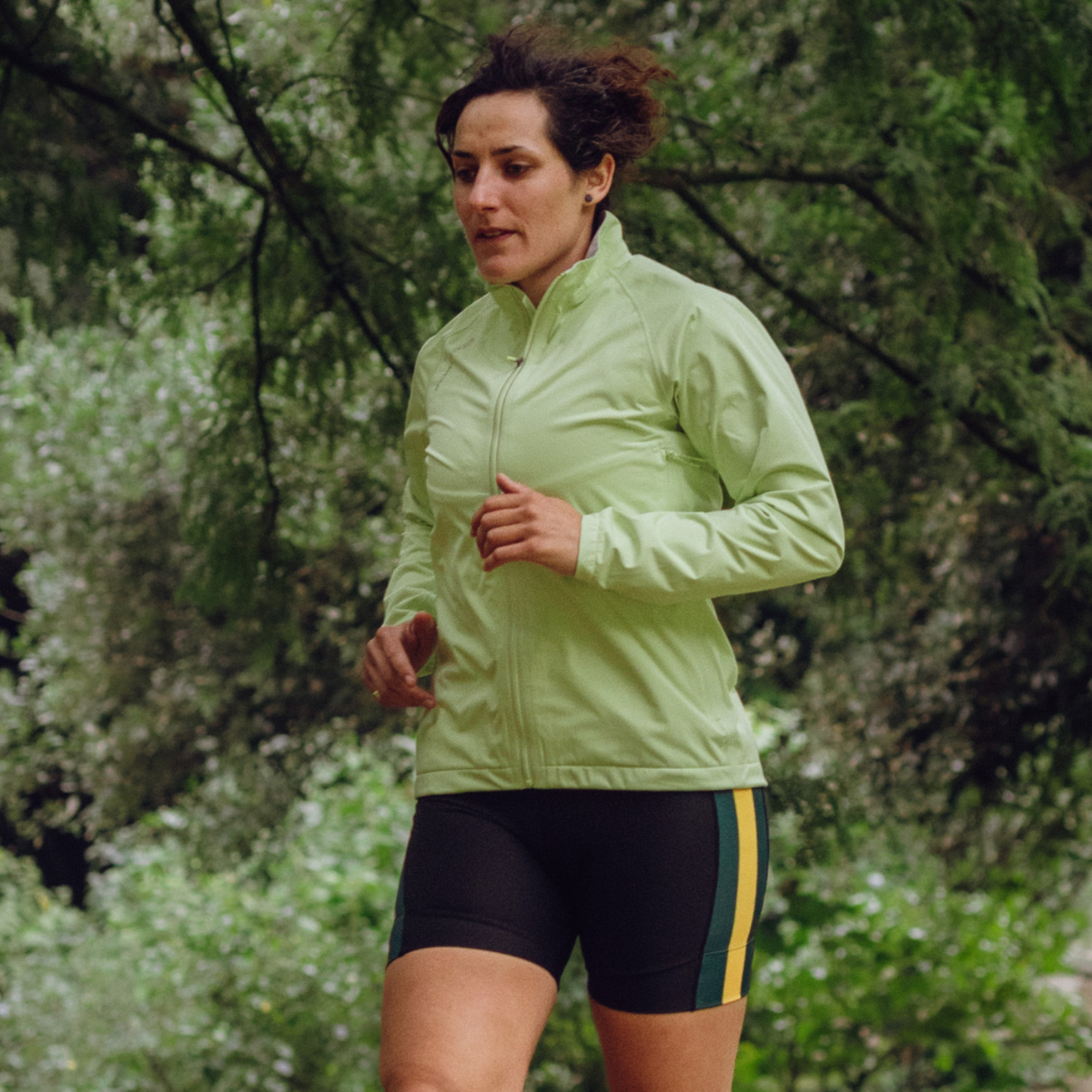 Woman running outdoors in a green Clourdburst jacket and black shorts with yellow accents.