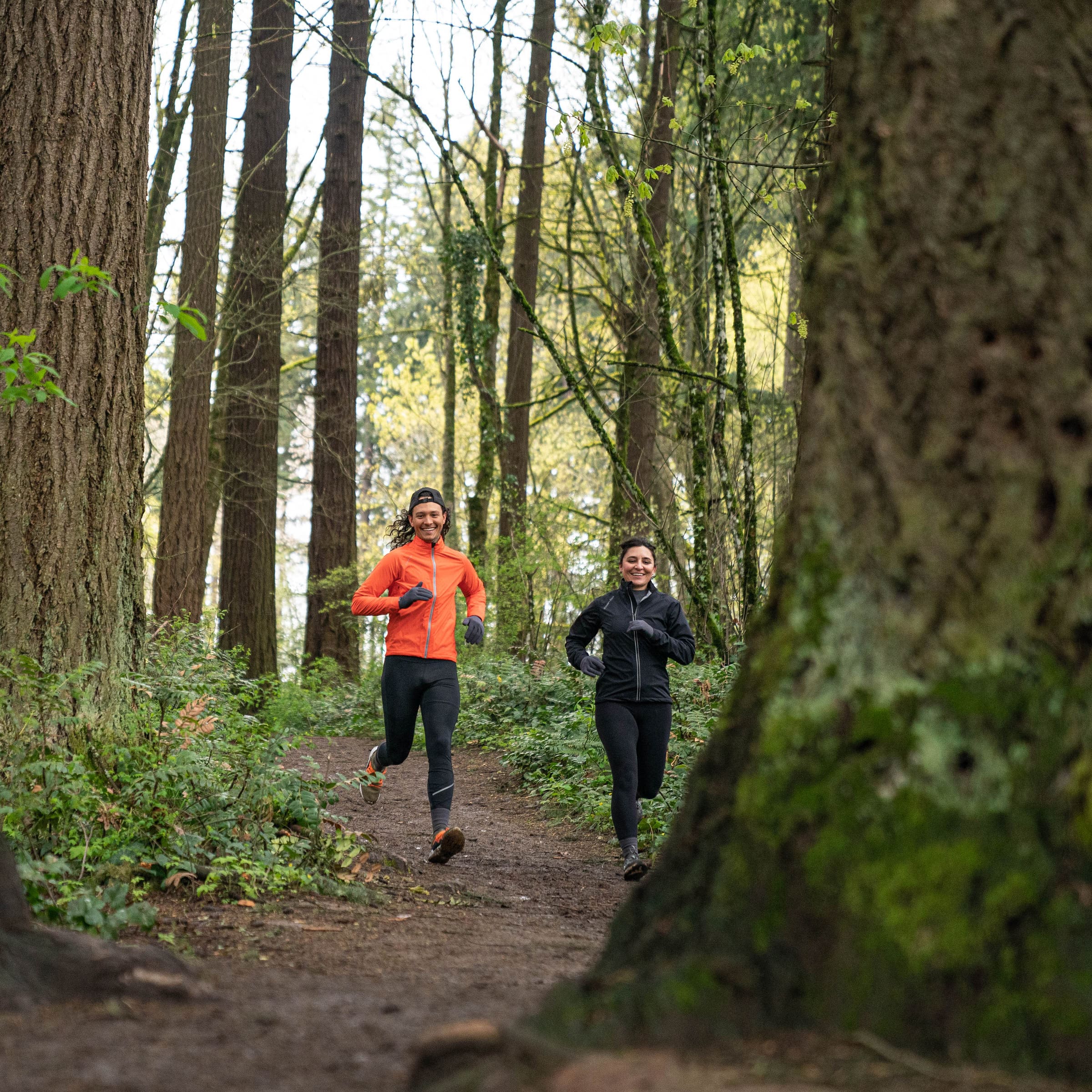 A man wearing an orange jacket and woman in a black jacket running along a trail path