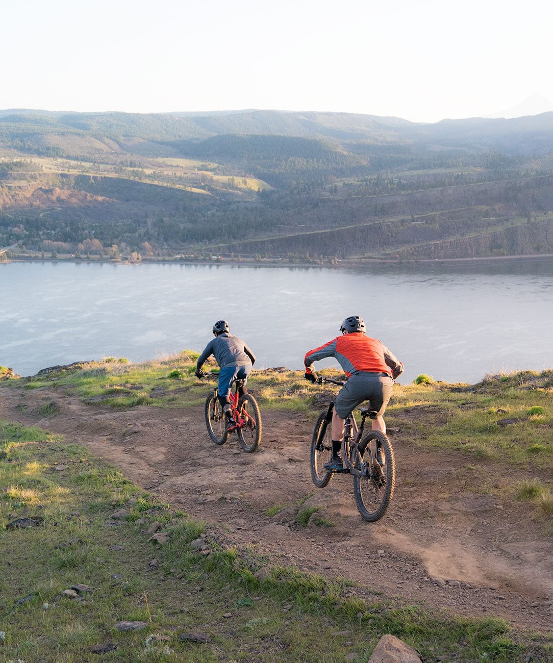 A scenic shot of two friends riding mountain bikes down a gravel path in the foreground, with a view of the rolling mountains in the background
