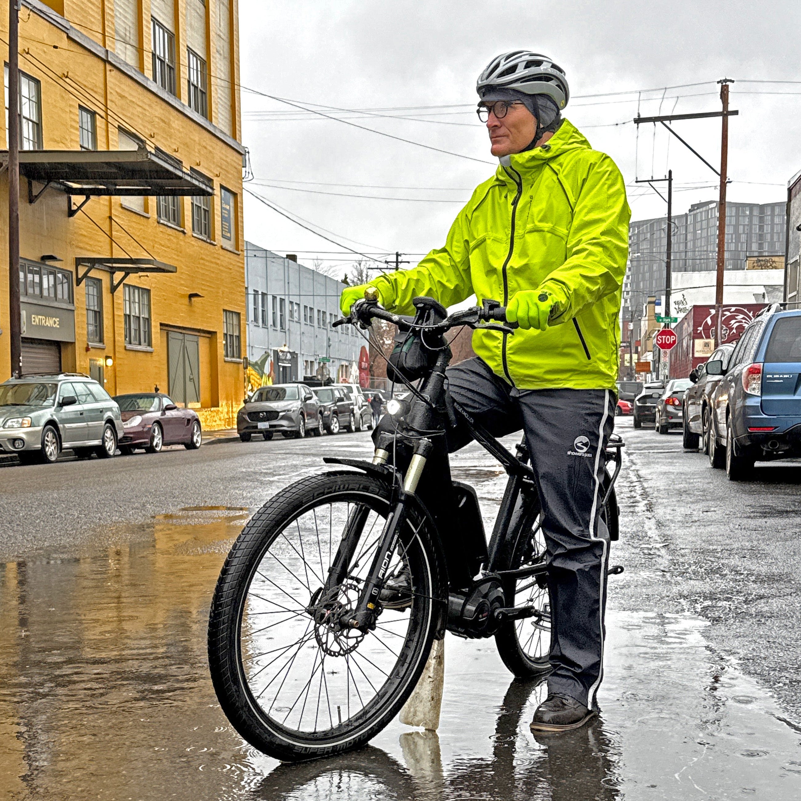 Person in a bright green Showers Pass jacket standing next to a bicycle on a city street with cars and buildings in the background.