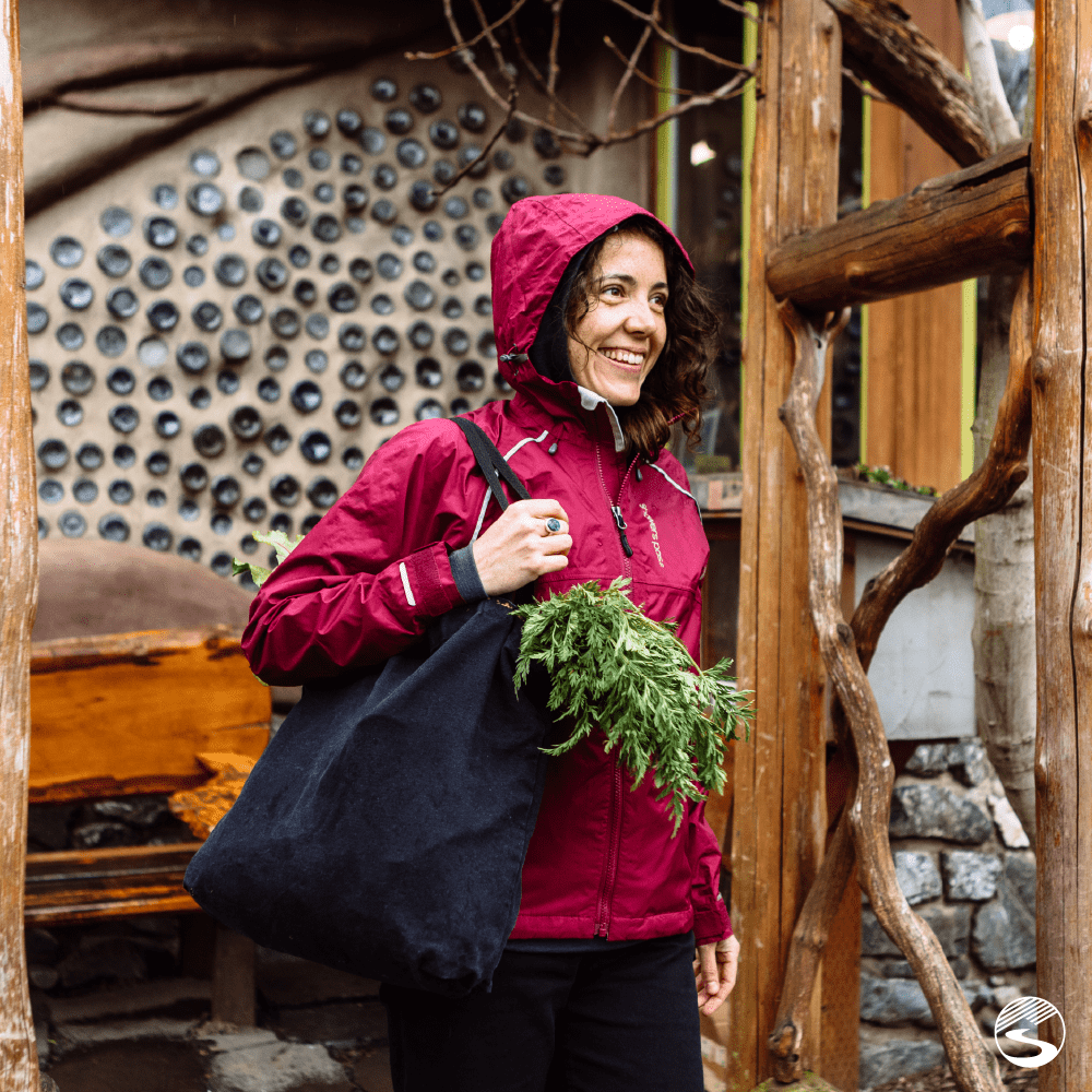A smiling woman wearing a ruby red-colored rain jacket by Showers Pass with a grocery bag slung over her shoulder