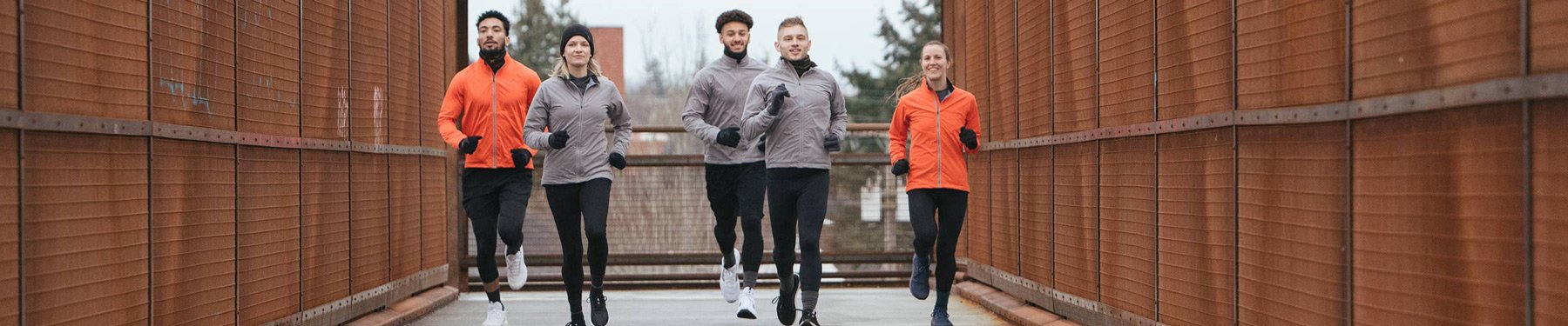 Five men and women wearing Showers Pass running apparel, jogging across a pedestrian bridge on a grey day.