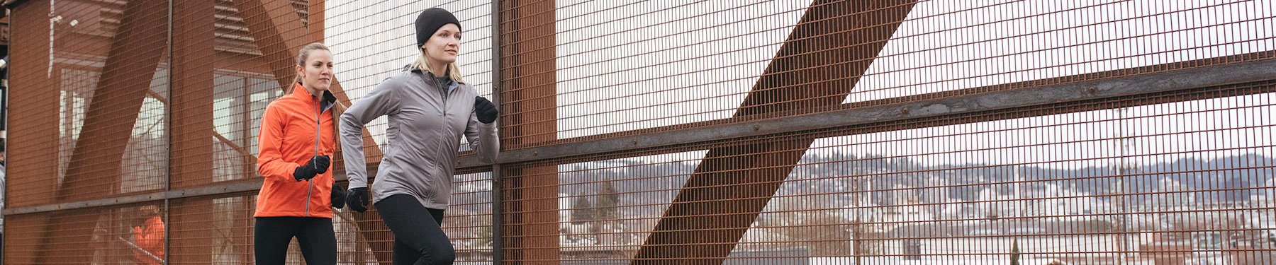 Two women wearing Showers Pass winter running gear, jogging across a bridge on a grey winter day.